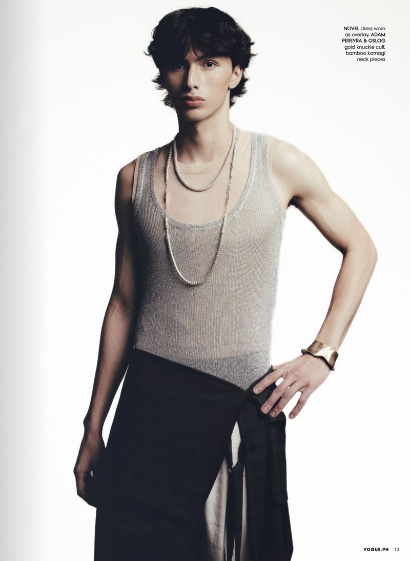 A slender, light-skinned adult male poses indoors wearing a gray tank top, dark skirt, and jewelry, against a white backdrop.