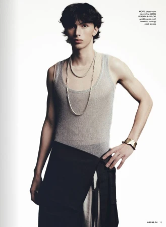 A slender, light-skinned adult male poses indoors wearing a gray tank top, dark skirt, and jewelry, against a white backdrop.