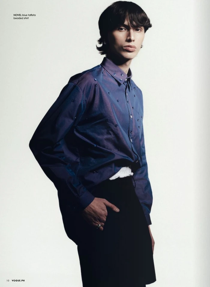 A male model posing in an iridescent blue beaded button-down shirt and black bottoms against a white studio background.