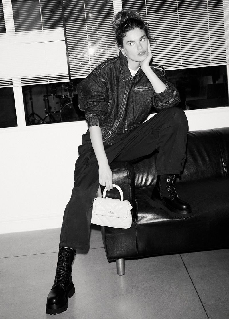A woman with a denim jacket and black pants sits on a black sofa, holding a light-colored purse, indoors with window blinds in the background.