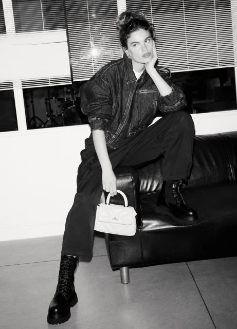 A woman with a denim jacket and black pants sits on a black sofa, holding a light-colored purse, indoors with window blinds in the background.