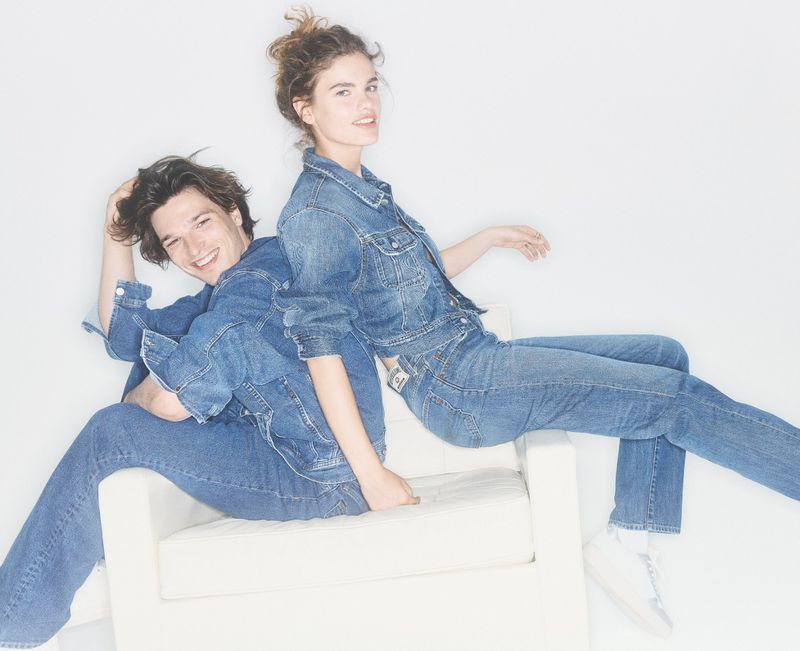Two young adults in denim jackets and jeans pose back-to-back on a white chair in a brightly lit studio, smiling at the camera.