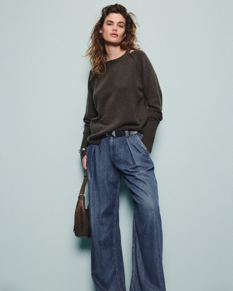 Fashion model poses in a studio wearing a dark green sweater, wide leg denim pants, and carrying a brown leather handbag.