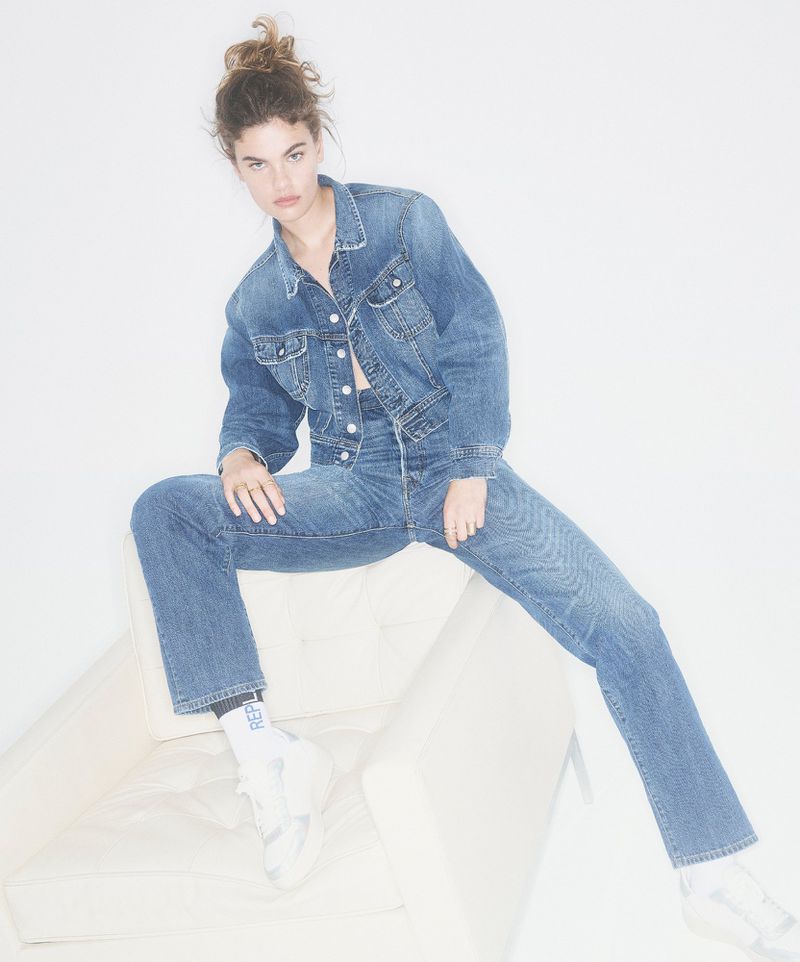 A young woman in a denim jacket and jeans poses on a white chair in a brightly lit photo studio, showcasing a casual and stylish look.
