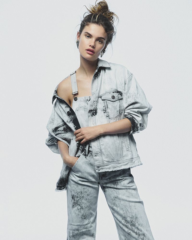Portrait of a woman in a light blue acid-wash denim jacket and matching pants standing in a photo studio against a white background.