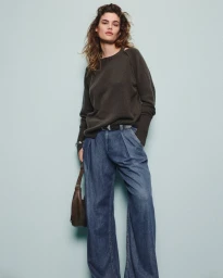 Model wearing a dark grey knit sweater and blue denim trousers holding a brown handbag against a seamless grey backdrop.