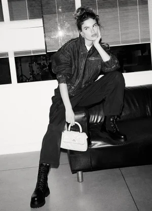 Model in denim jacket and dark trousers sitting on a leather sofa holding a light handbag in a monochrome fashion campaign shot