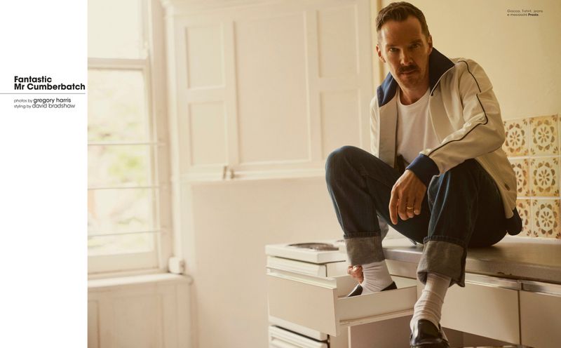 Adult male model sitting on a kitchen counter wearing a white jacket, jeans, white socks and black shoes in a bright, natural light indoor setting.