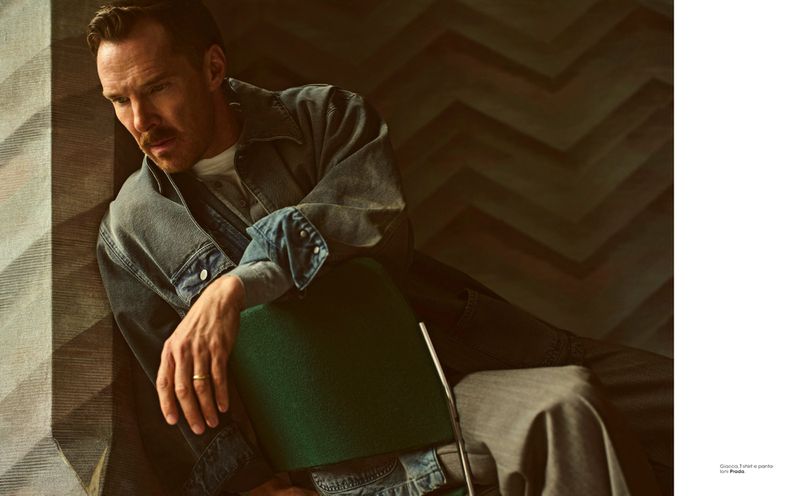 Benedict Cumberbatch, an adult male, is pictured indoors, seated on a green chair, wearing a denim jacket and grey pants in a medium shot.