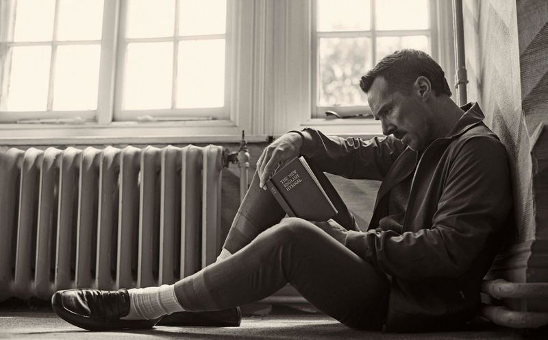 Monochrome image of an adult male sitting on the floor reading a book by a radiator and a window in a well-lit indoor room.