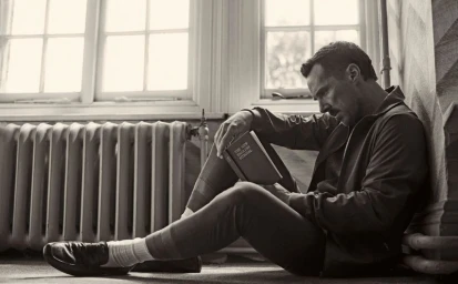 Monochrome portrait of Benedict Cumberbatch seated on the floor reading a book against a windowed wall.