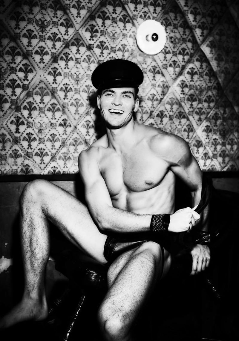 Black and white portrait of muscular adult male wearing cap in dark interior with patterned wallpaper and shiny vinyl seat.