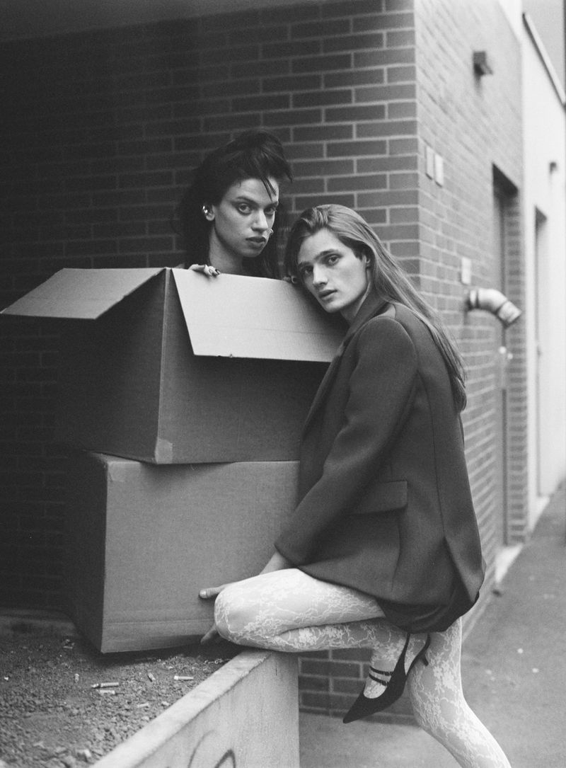 Two women pose with cardboard boxes against a brick wall in a black and white fashion shot featuring textured tights and a blazer.