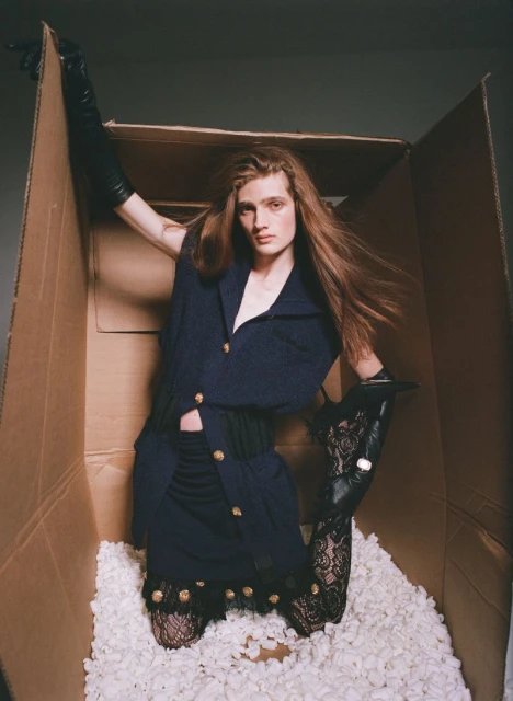 A woman with long hair wearing a navy-blue dress and black lace gloves inside a cardboard box filled with white packing peanuts.
