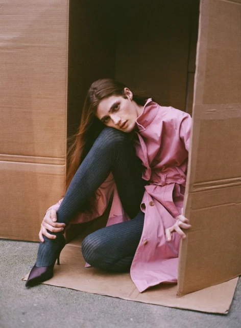 A person with long brown hair sitting inside a cardboard box wearing a pink jacket, dark ribbed tights, and dark high-heeled shoes.