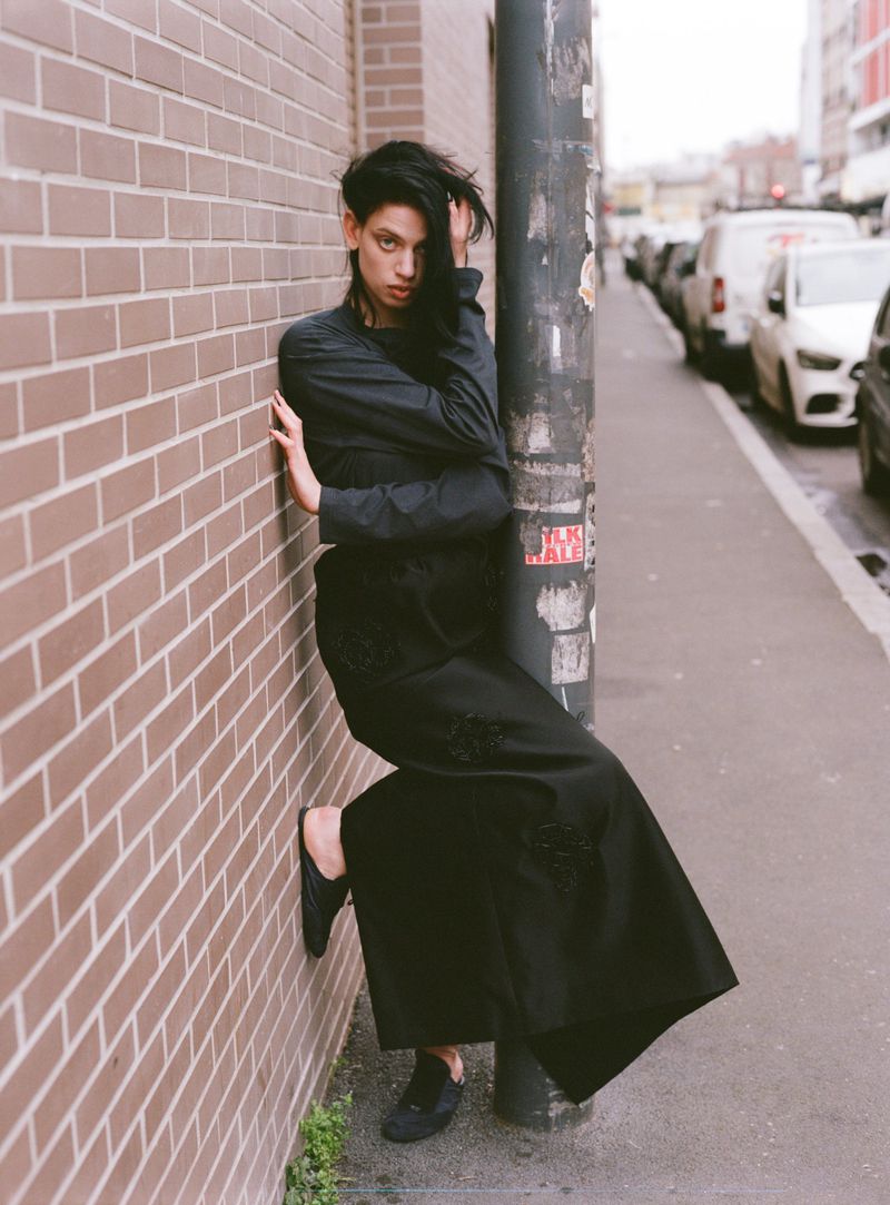 A stylish young woman poses against a brick wall on a city street, wearing a black dress and flats, looking directly at the camera.