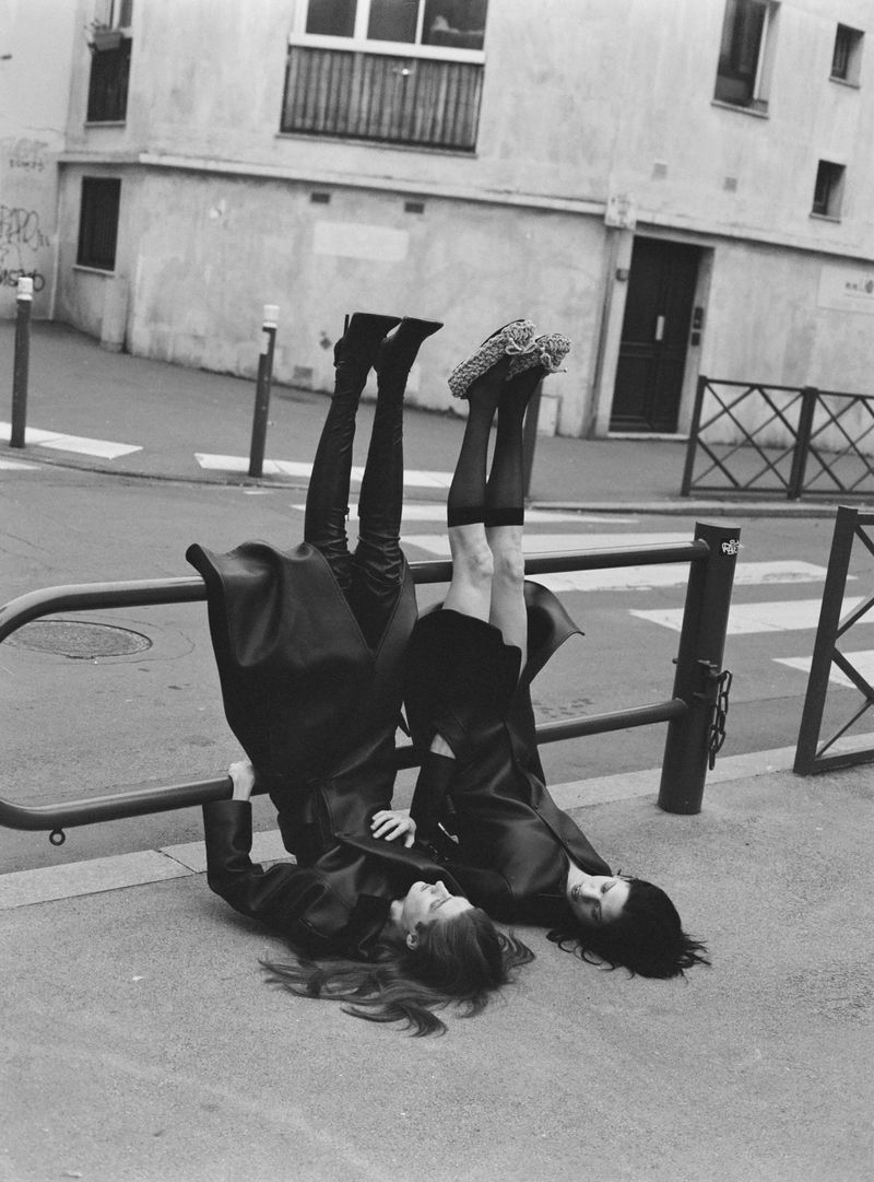 Two women in black outfits are playfully posed upside down against a metal railing in an urban setting, showcasing a monochrome aesthetic.
