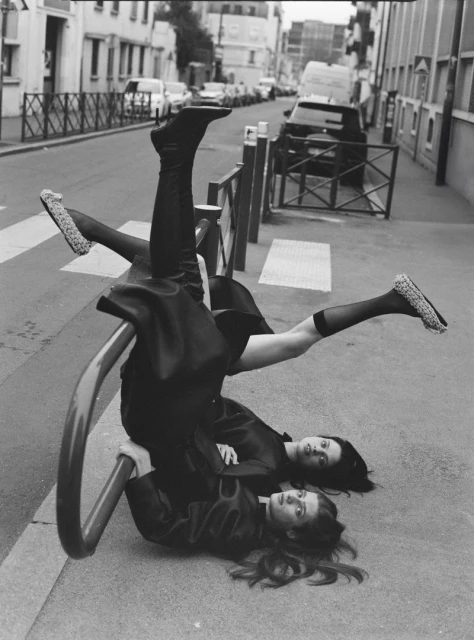 Two fashion models strike a pose lying on the street with one model inverted on a metal barrier, capturing a unique urban style.