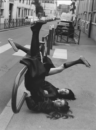 Two fashion models strike a pose lying on the street with one model inverted on a metal barrier, capturing a unique urban style.