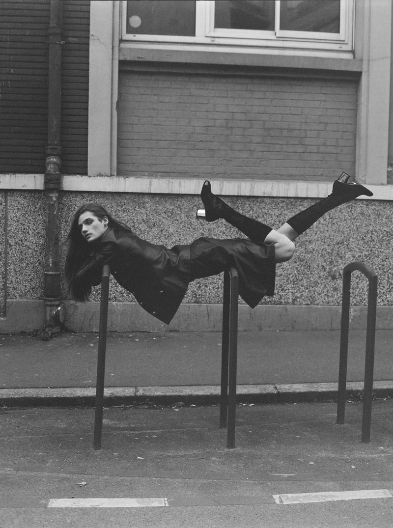 Monochrome shot of a woman in a coat and boots lying on a metal structure in an urban environment, creating an editorial fashion image.