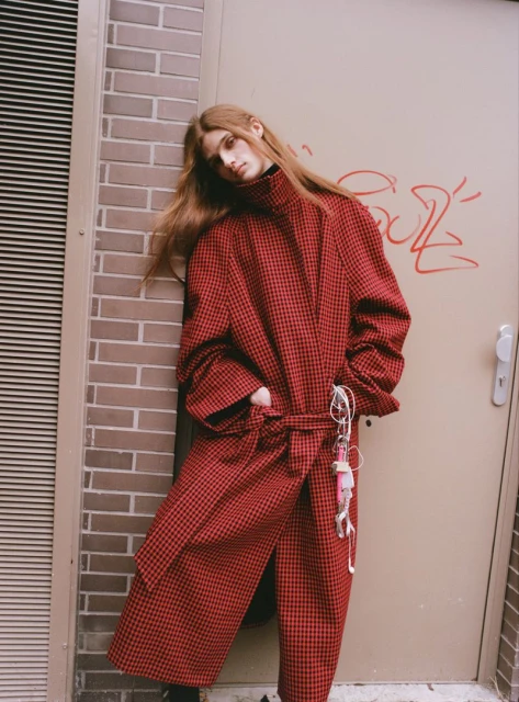 Redheaded woman in a long, belted red and black checkered coat leans against a brick wall with graffiti; fashion editorial shoot.