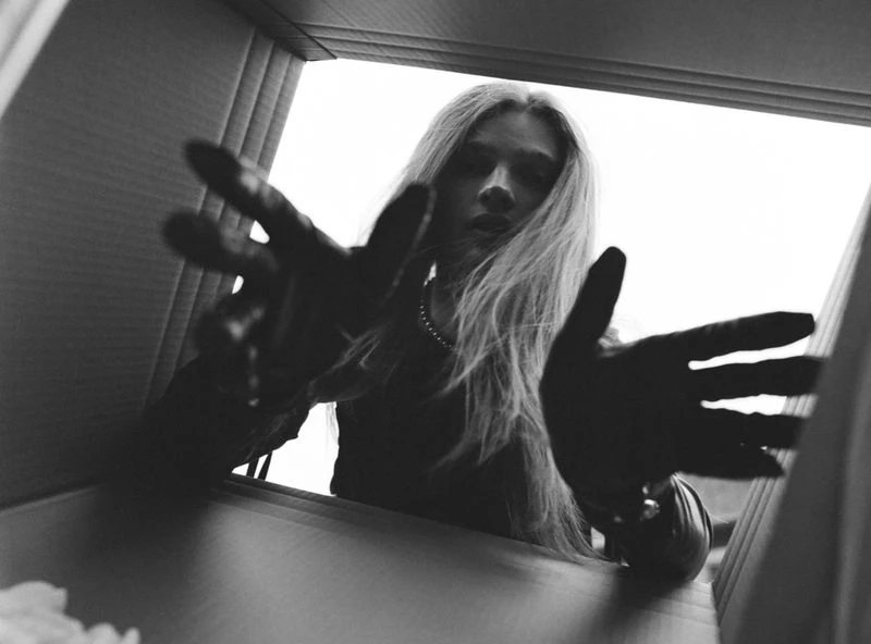 Monochrome portrait of a young adult woman with long blonde hair reaching inside a cardboard box from a low-angle perspective.