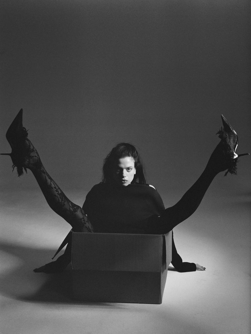 A stylish woman sits in a box with legs extending upward, showcasing heels in a striking black and white fashion photograph.