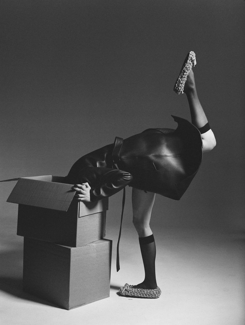 Artistic black and white image of a woman in a black coat emerging from a cardboard box wearing woven footwear with one leg raised.