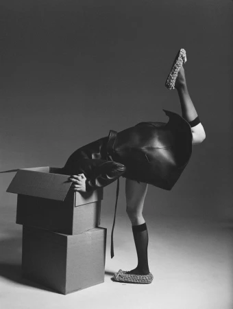 Artistic black and white image of a woman in a black coat emerging from a cardboard box wearing woven footwear with one leg raised.