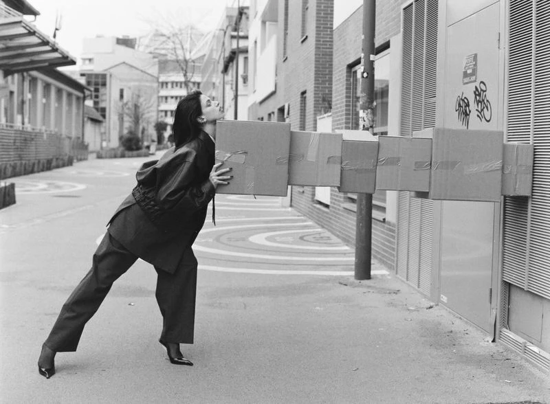 Woman in suit struggles to carry a tower of taped cardboard boxes on a city sidewalk with buildings in the background, monochrome.