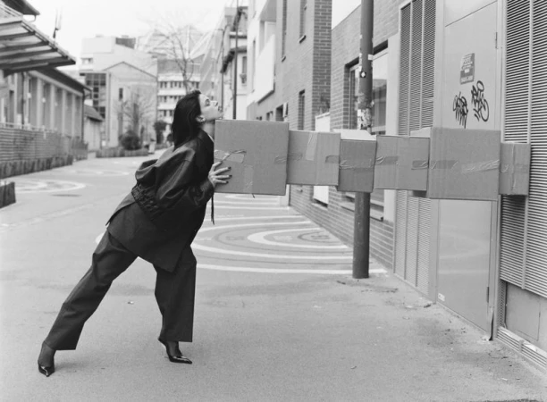 Woman in suit struggles to carry a tower of taped cardboard boxes on a city sidewalk with buildings in the background, monochrome.