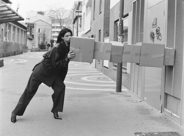 Woman carrying a train of cardboard boxes down a city street with apartment buildings in the background, black and white photography.