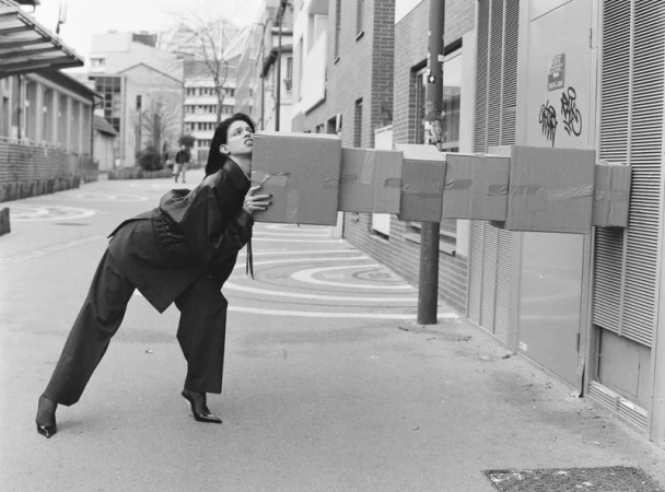 Black and white image of a woman in a pantsuit holding a series of taped cardboard boxes on a city street with buildings in the background.