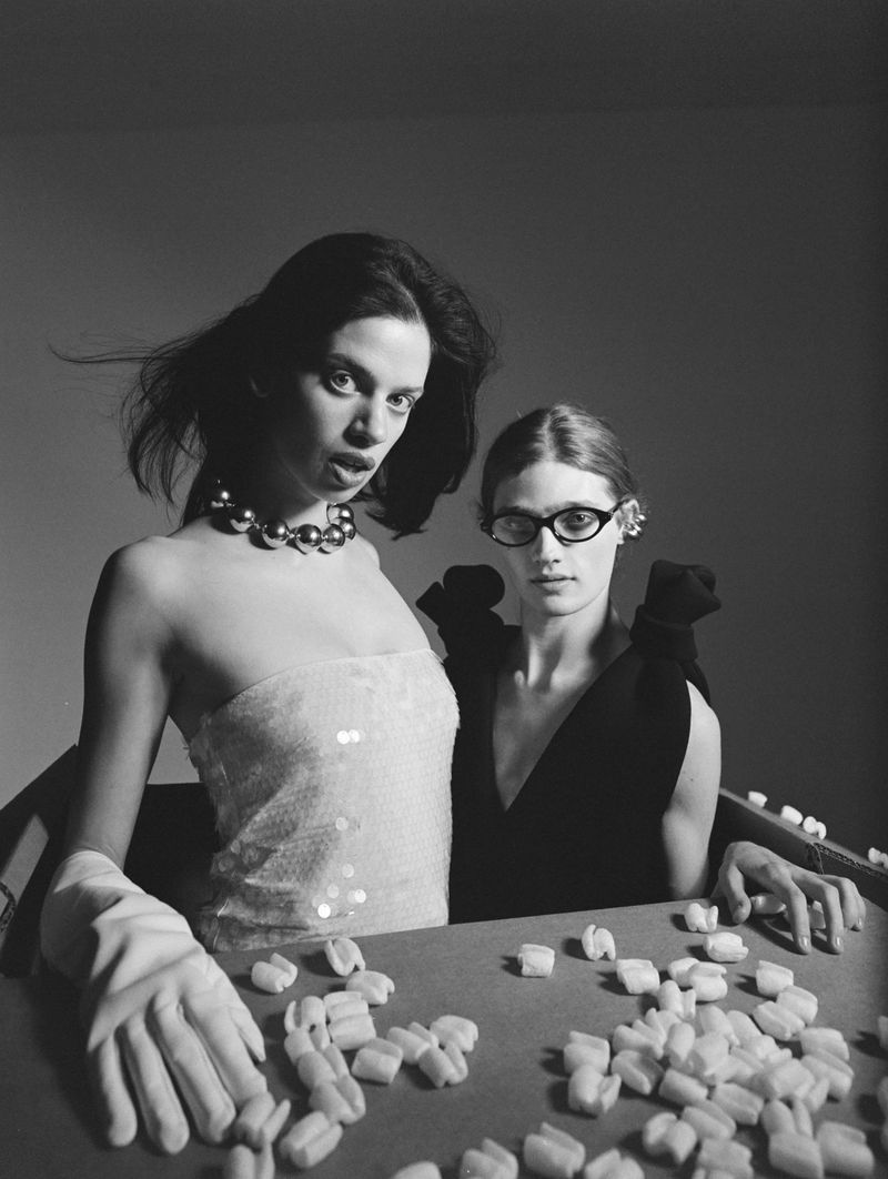 Two women pose in a monochrome image, one wearing a strapless top and necklace, the other glasses, in a studio setting with packing peanuts.