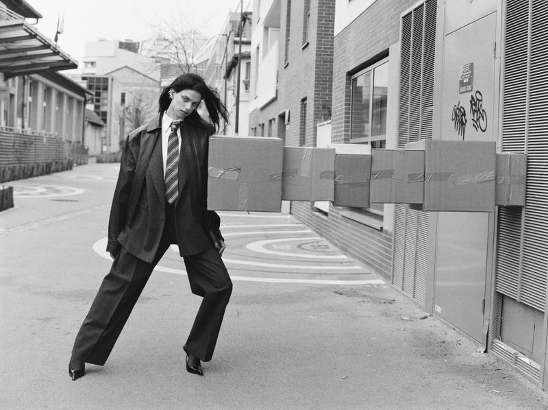 Monochrome fashion shot of a woman in an oversized suit, balancing a stack of cardboard boxes on her shoulder on a city street.