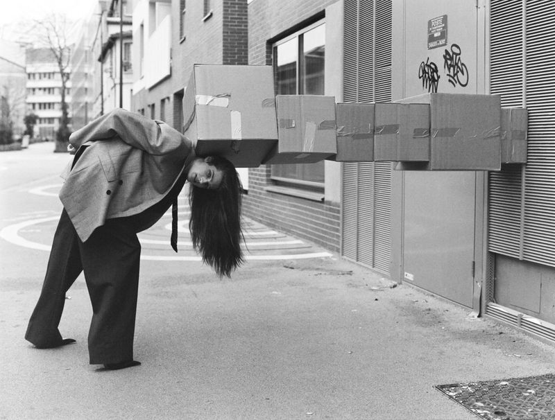 Young woman in grey jacket balances a stack of taped cardboard boxes on her neck outdoors in a street in a black and white photograph.