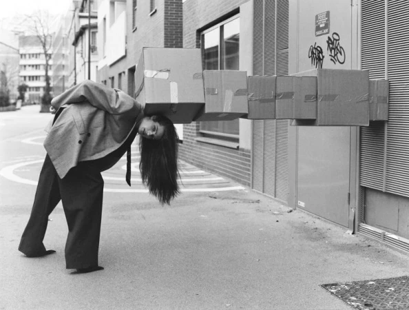 Young woman in grey jacket balances a stack of taped cardboard boxes on her neck outdoors in a street in a black and white photograph.