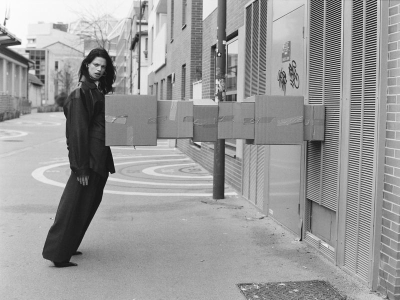 Monochrome fashion shot of a person in dark clothing standing outside near brick buildings, holding a horizontal series of cardboard boxes.