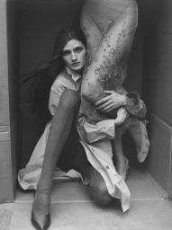 Model Lily McMenamy seated inside a box wearing a trench coat and tights, holding onto embellished sheer dress legs.