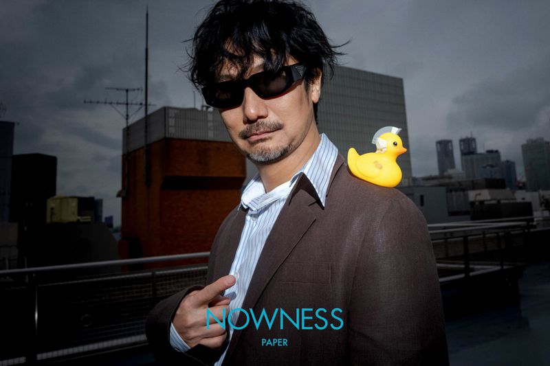 Man in a suit stands on a rooftop with a toy duck on his shoulder, city buildings and a cloudy sky in the background.