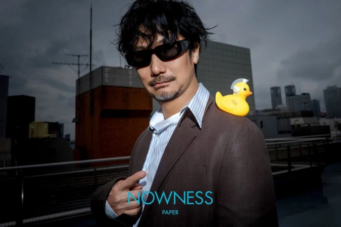 Man in a suit stands on a rooftop with a toy duck on his shoulder, city buildings and a cloudy sky in the background.