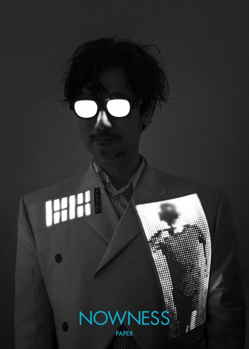 Portrait of a person wearing a suit and sunglasses, with the words Nowness Paper visible. The person's sunglasses are glowing.