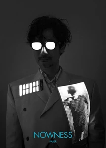 Portrait of Hideo Kojima wearing a light suit with glowing digital panels and illuminated glasses against a dark background