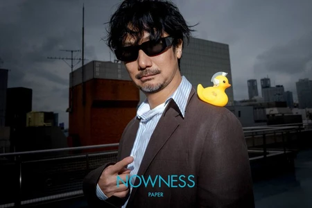Hideo Kojima wearing sunglasses and a brown blazer with a small rubber duck on his shoulder outdoors against a city skyline