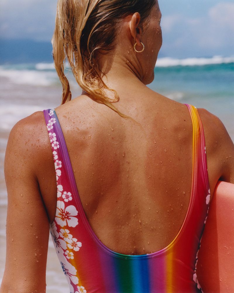 Woman in a swimsuit standing on a beach with a bodyboard. Rainbow colored swimsuit with flower details and fair skin visible.