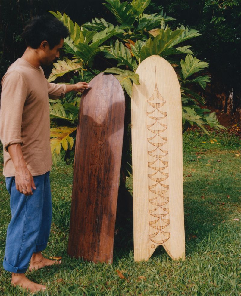Man stands beside two wooden surfboards in a grassy outdoor setting with lush green foliage. One surfboard is light, the other dark.