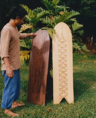 Man stands beside two wooden surfboards in a grassy outdoor setting with lush green foliage. One surfboard is light, the other dark.
