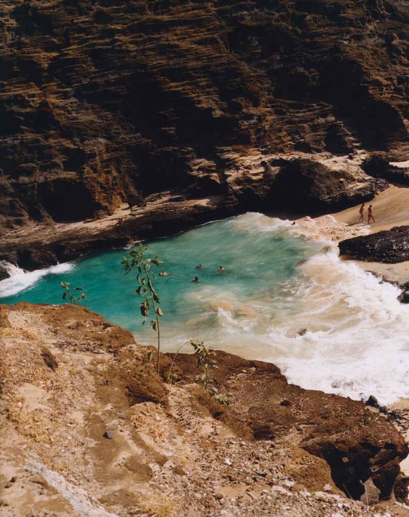Aerial view of turquoise ocean water in a rocky cove with people swimming and two figures walking on a sandy beach.