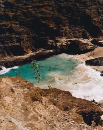 Aerial view of turquoise ocean water in a rocky cove with people swimming and two figures walking on a sandy beach.