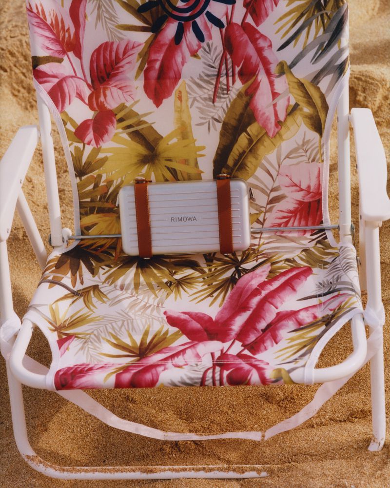 Floral beach chair on sand with a Rimowa suitcase secured on top with brown straps, showcasing travel and leisure.